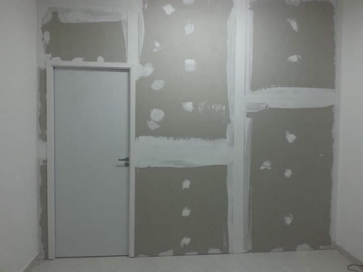 uma parede interna com gesso e juntas de tinta branca, com uma porta de cor branca a esquerda  — texto:  