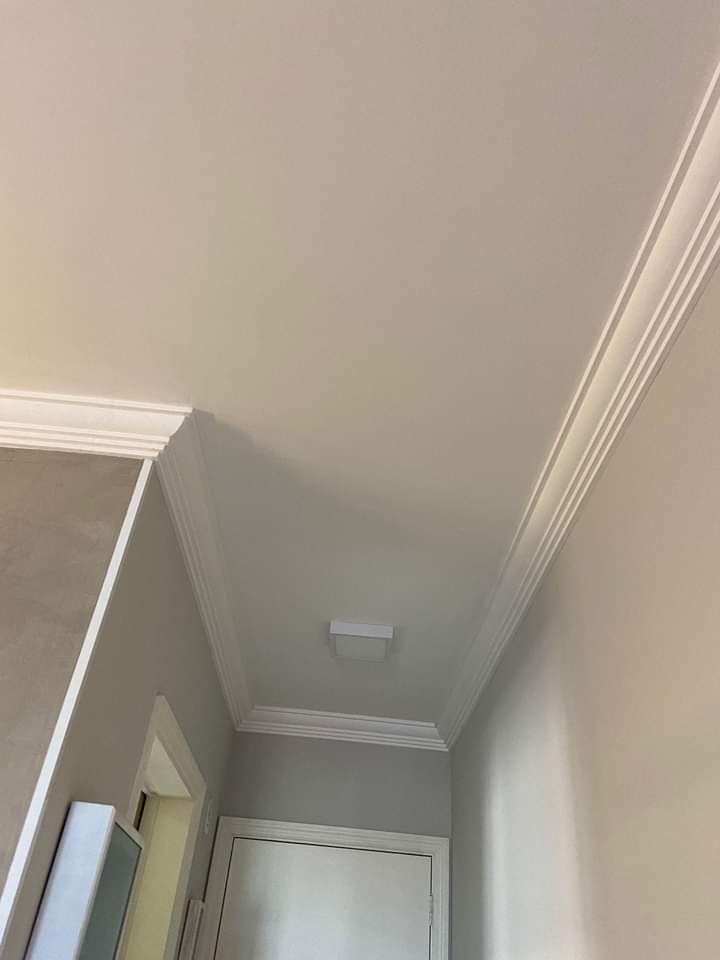 parte superior de um corredor interno com teto branco, molduras de gesso e uma porta em fundo  — texto:  
